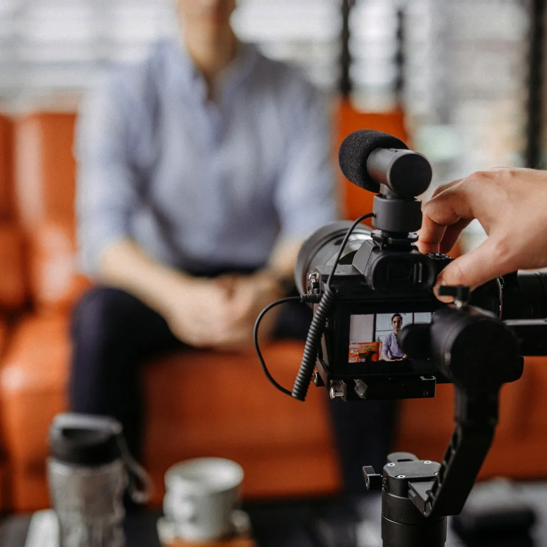 A camera on a tripod with a shotgun microphone, filming a seated person who is out of focus behind it.