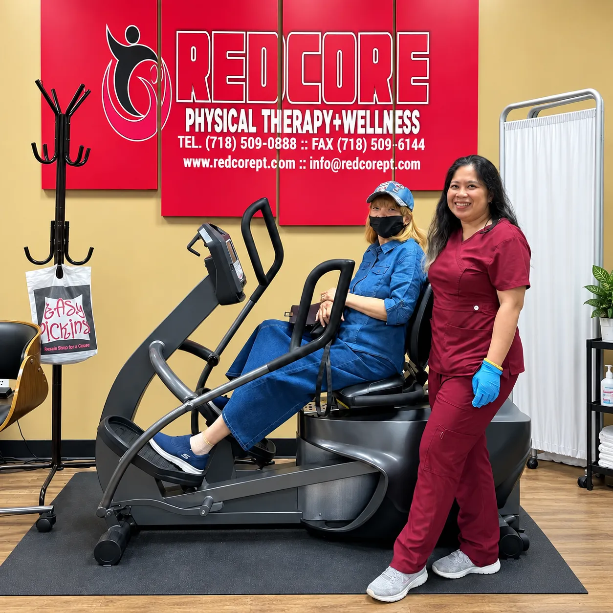 Un terapeuta de RedCore con uniforme granate, de pie junto a un paciente que usa una elíptica con asiento, bajo el letrero de RedCore.