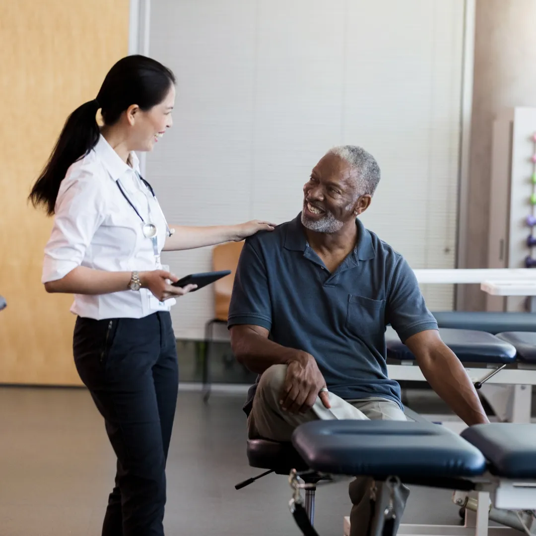 A clinician and an older patient smiling and holding hands mid-conversation in a bright communal room.